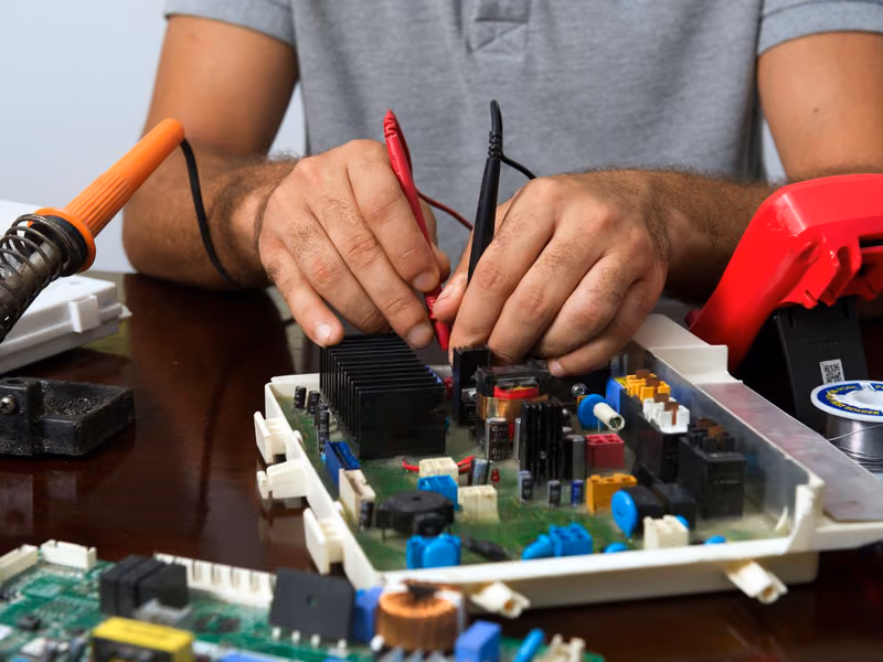 Hands probing a circuit board with a multimeter while working at a bench